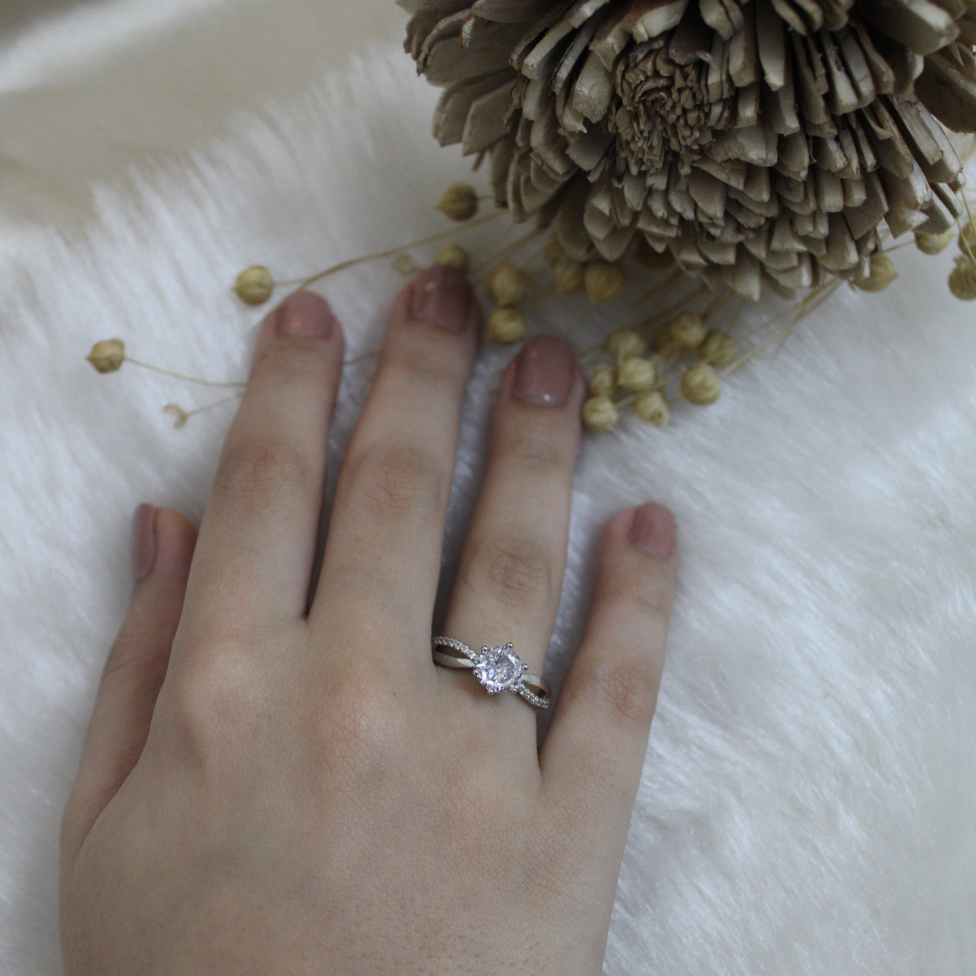 Hand wearing a ring with a diamond, surrounded by small yellow flowers and a pine cone on a white surface.