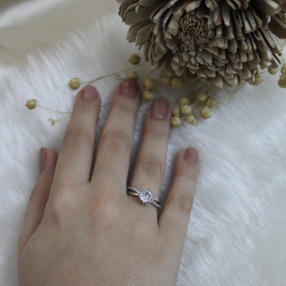 Hand wearing a ring with a diamond, surrounded by small yellow flowers and a pine cone on a white surface.