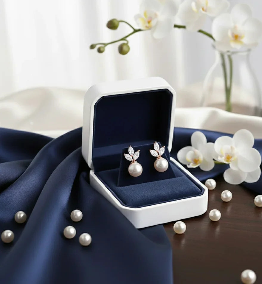 Elegant pearl earrings with diamond accents in a navy velvet box, surrounded by white orchids and loose pearls