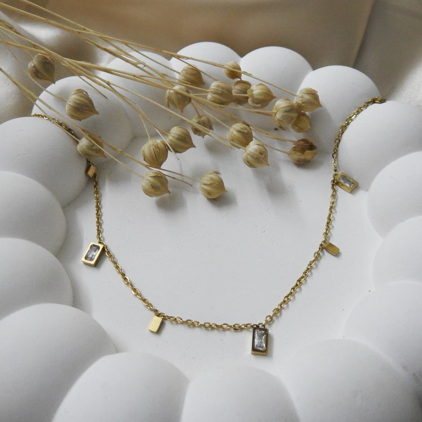 Gold necklace with small rectangular pendants on a white surface with dried flowers.