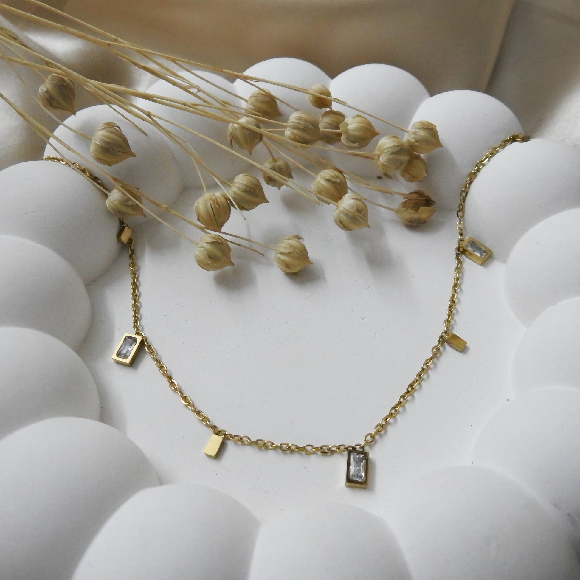 Gold necklace with small rectangular pendants on a white surface with dried flowers.